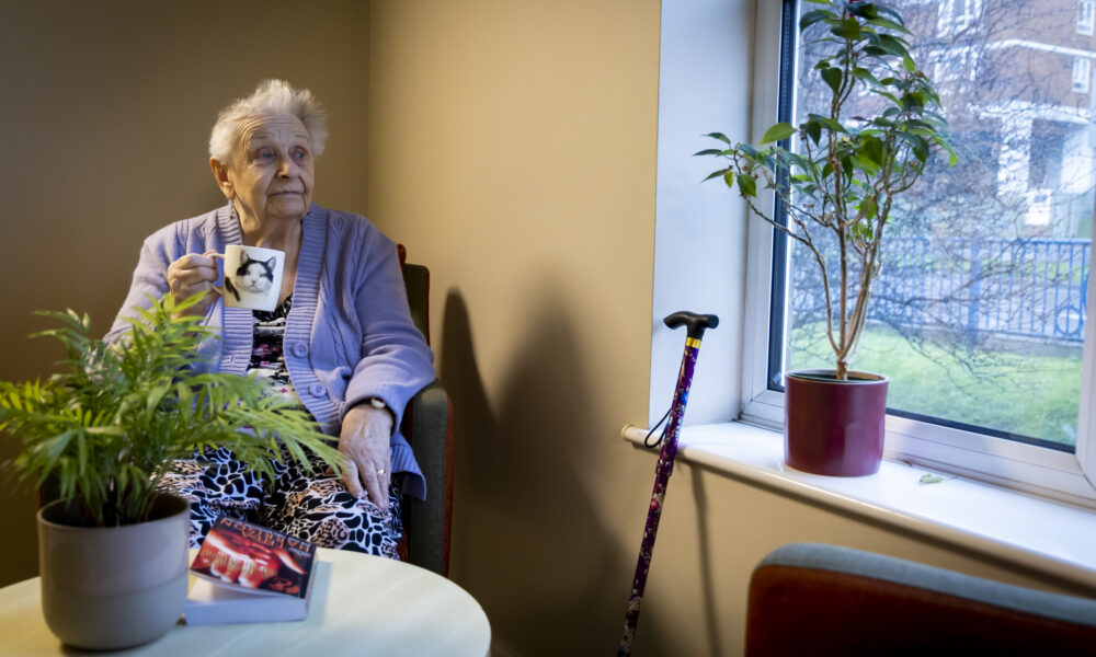 Resident Rights Month. A woman with short white hair holding a cat mug looks out a window. A houseplant is on the table in the foreground and a cane rests against the window to her side.