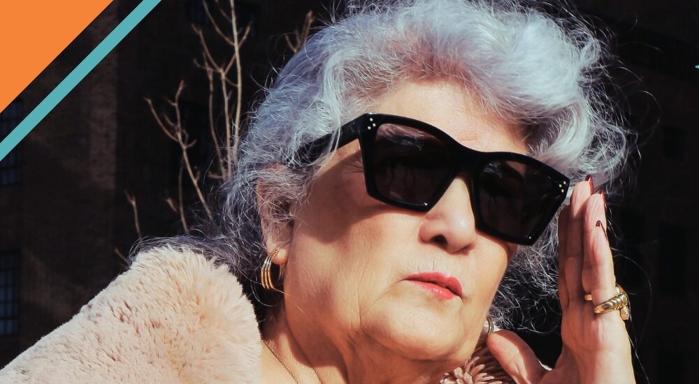 A woman with white hair touches her black sunglasses. She is wearing a fur coat and gold jewelry